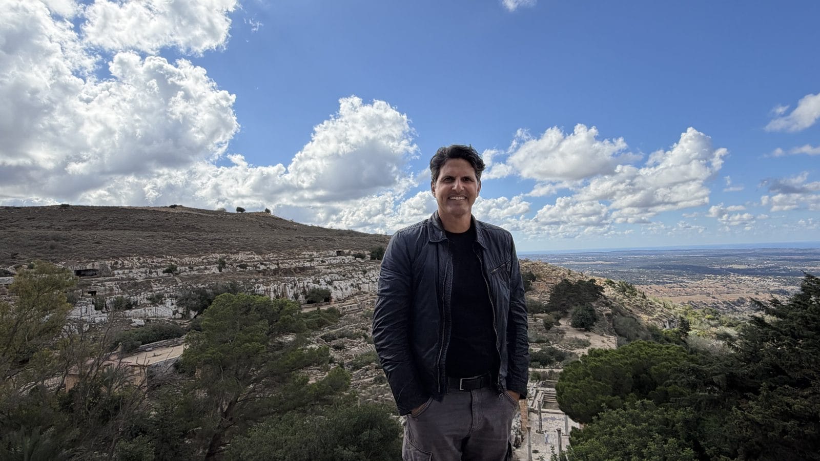 A man stands on a rocky hillside overlooking a sprawling valley under a blue sky with clouds.