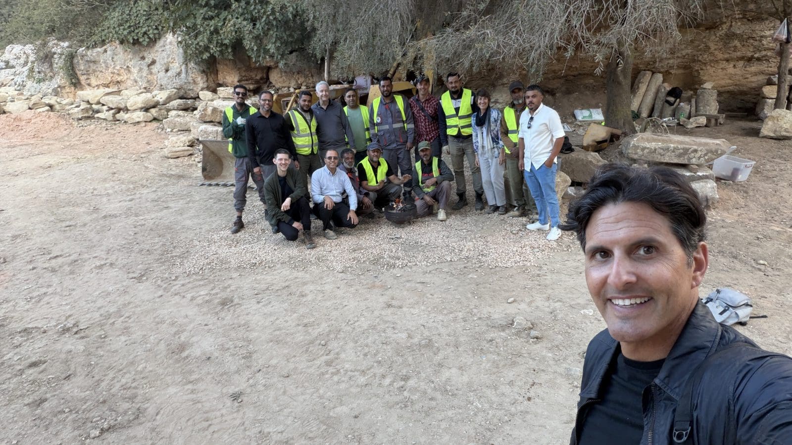 A man takes a selfie in the foreground with a large group of people standing in front of a rocky cliffside, many wearing high-visibility safety vests.