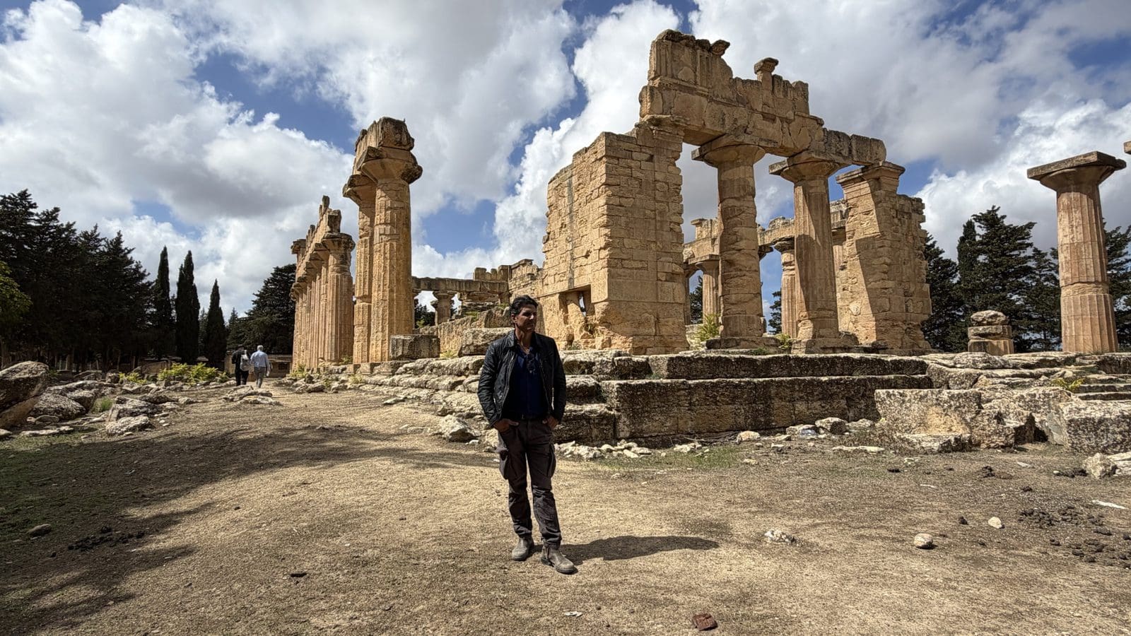 A man stands in front of the ancient stone ruins of a Greek temple under a cloudy sky.