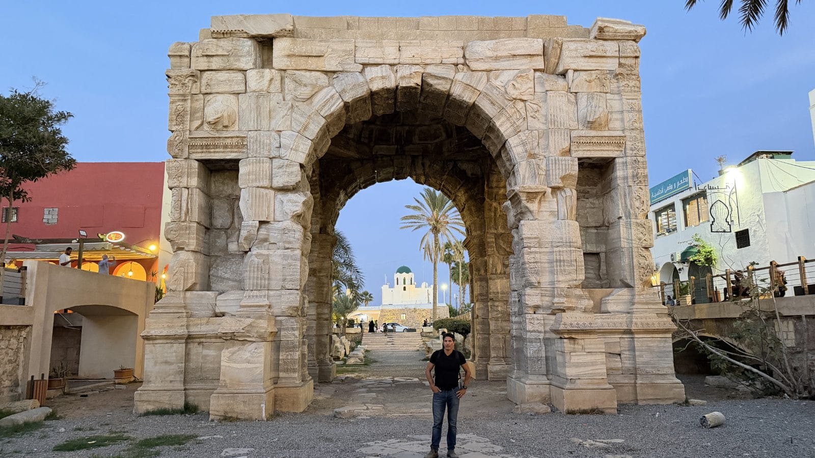 A man stands in front of a large, ancient stone archway with a white domed building visible through the center.