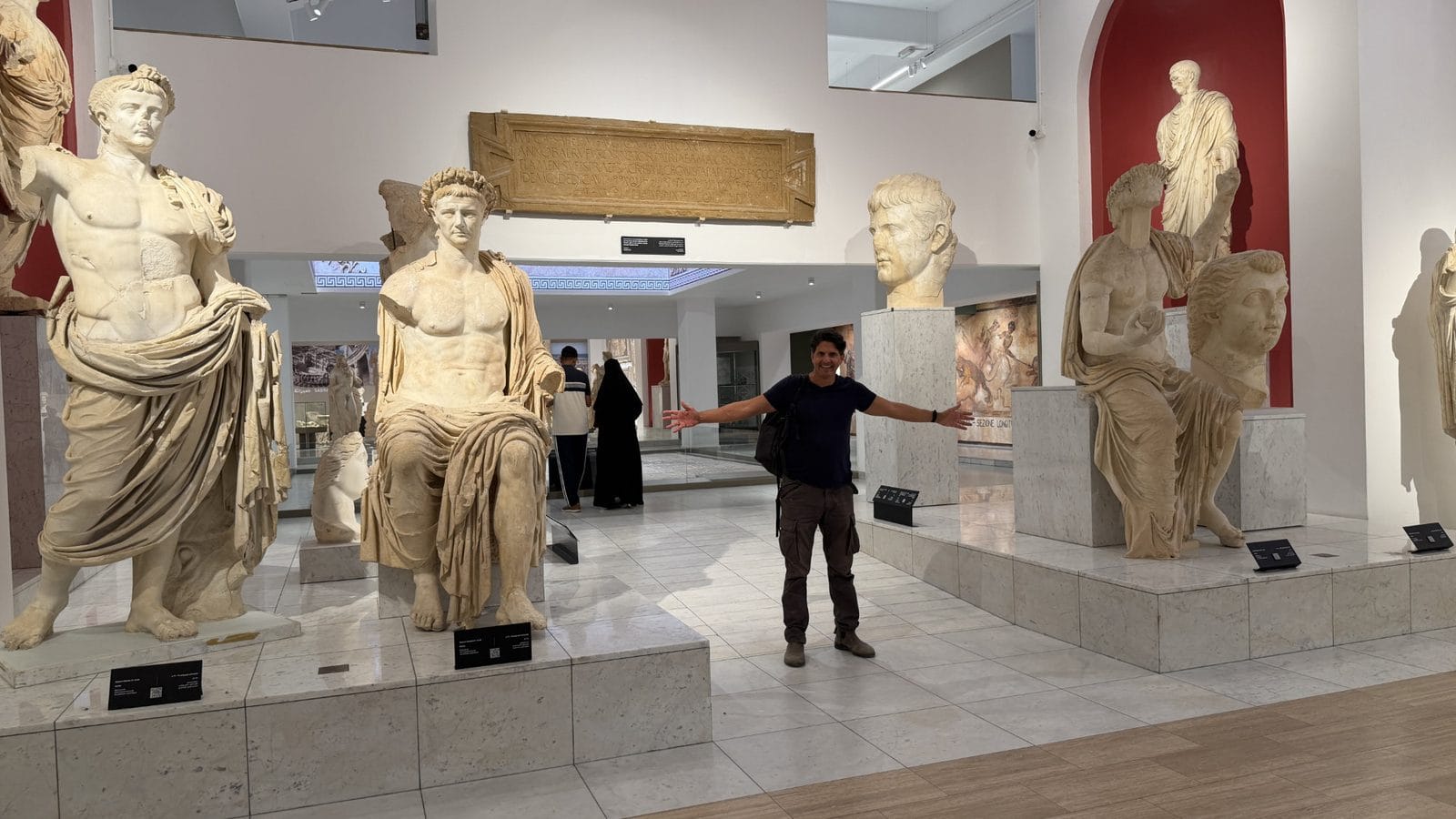 A person stands with arms outstretched in a museum gallery among several marble Roman statues and a large inscribed stone tablet.
