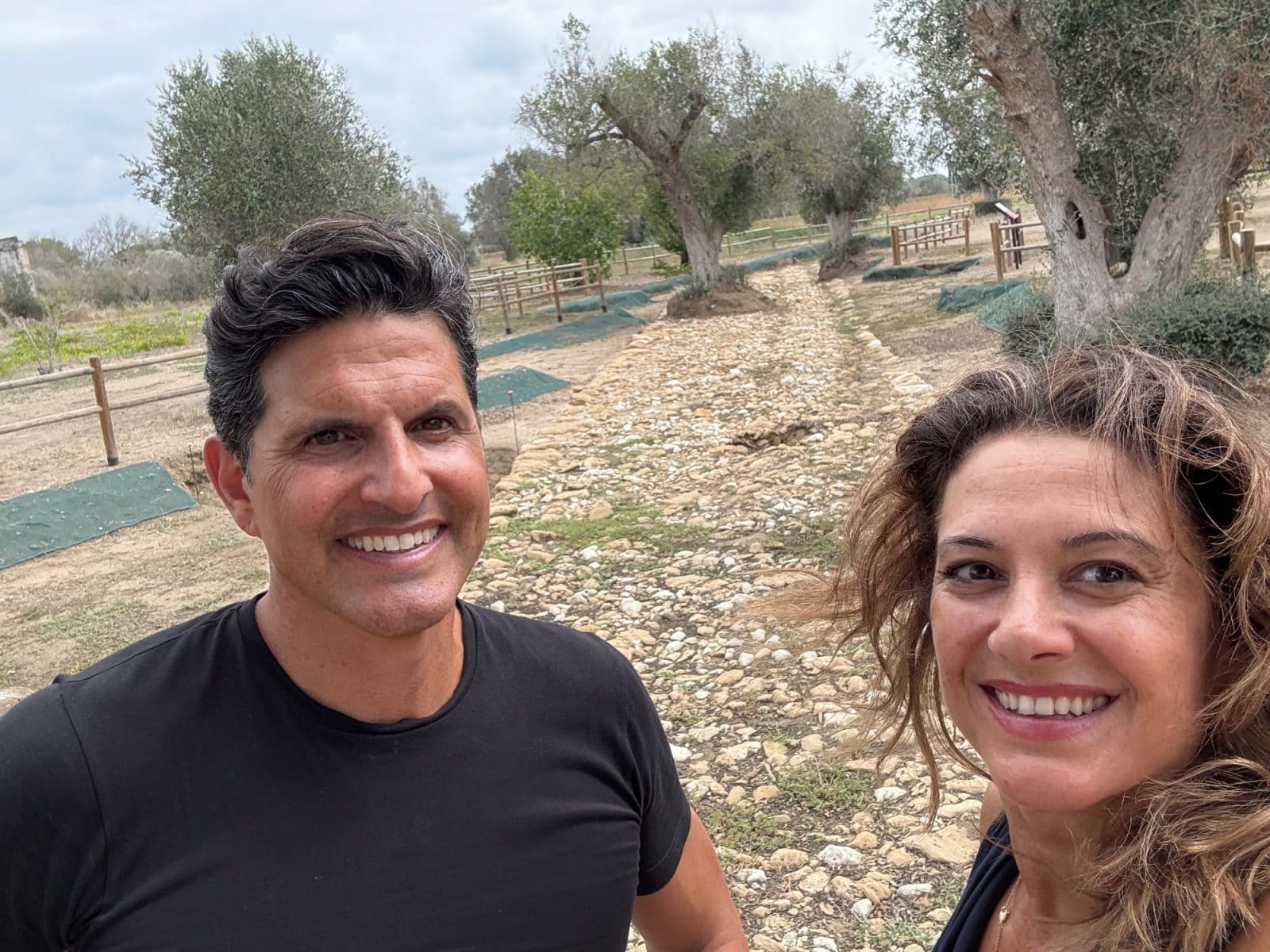 A man and a woman smiling for a selfie on a stone path in an olive grove.