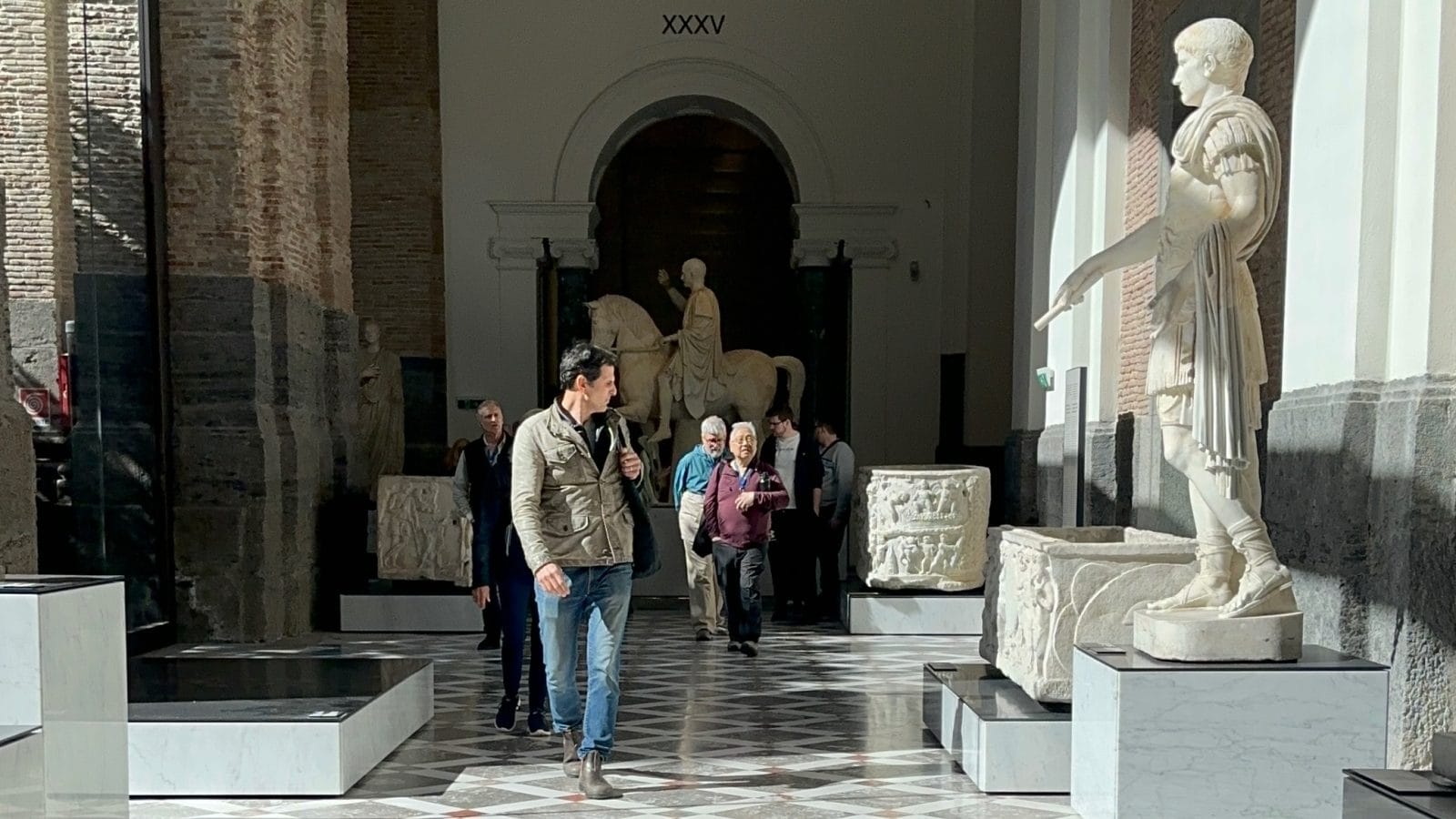 Visitors walk through a museum gallery featuring ancient Roman statues, including a prominent marble statue of a man in armor on the right.
