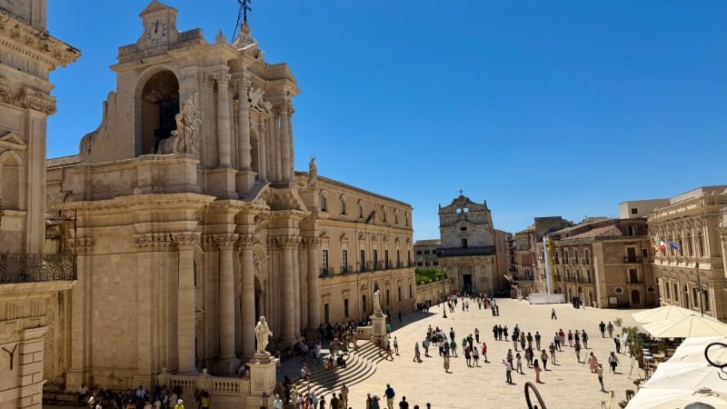 The Syracuse Cathedral, a large Baroque church with columns and statues, overlooking a sunlit square filled with pedestrians.