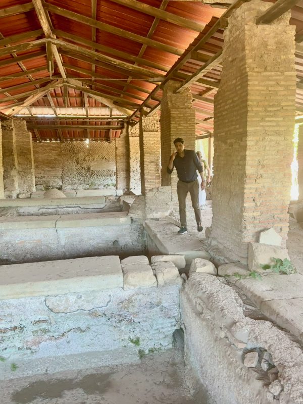 A person stands among ancient stone ruins under a red-roofed structure with brick pillars and partially exposed foundations.