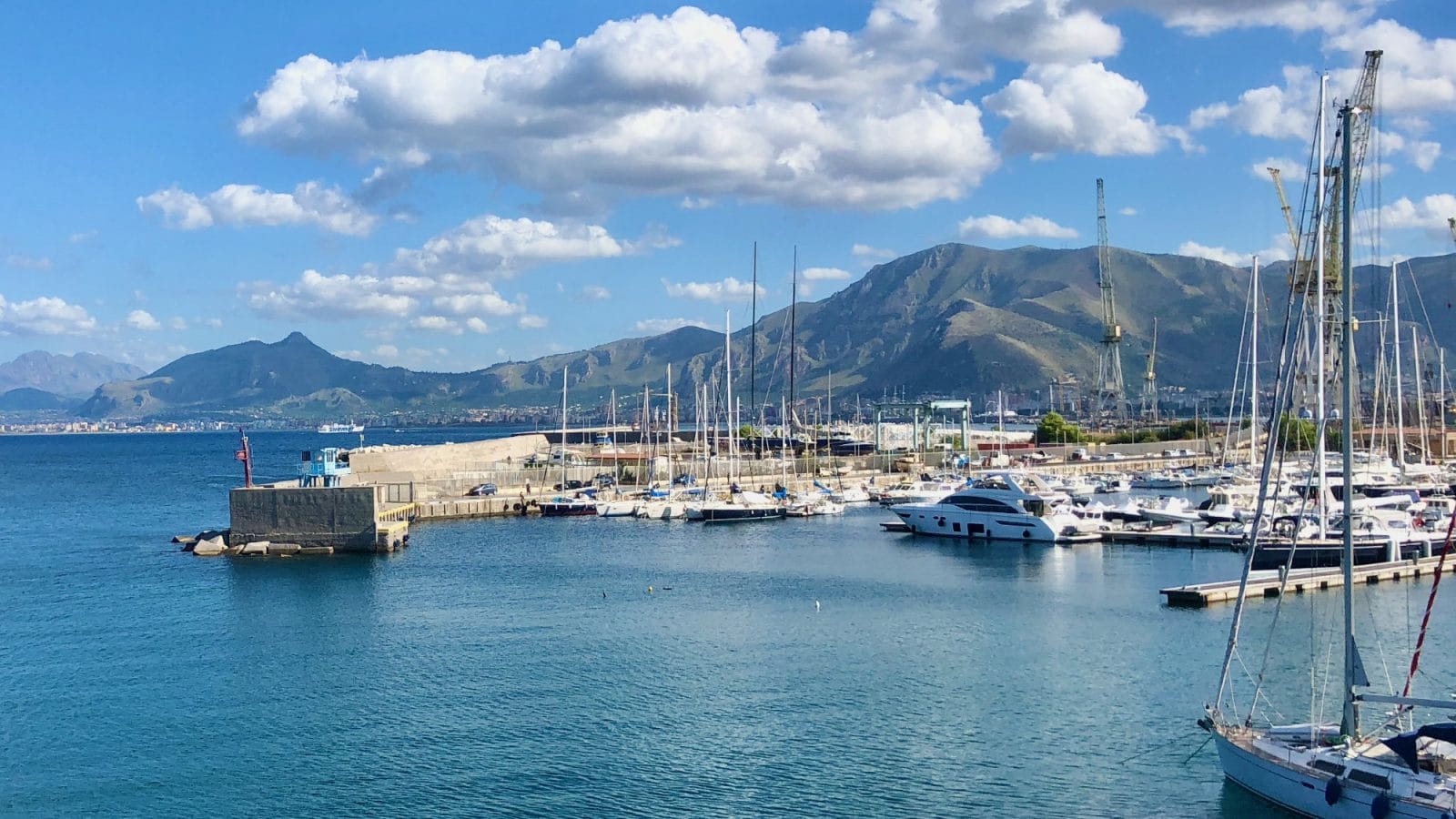 A marina with yachts and sailboats docked in front of mountains under a blue sky with clouds.