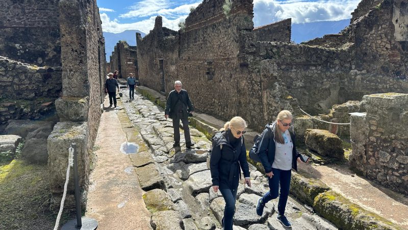 Tourists walk along a narrow, uneven stone street between ancient stone ruins in Pompeii.