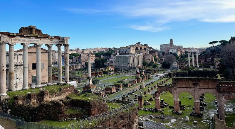 The Roman Forum, featuring ancient stone columns, ruins, and grassy pathways under a clear blue sky.
