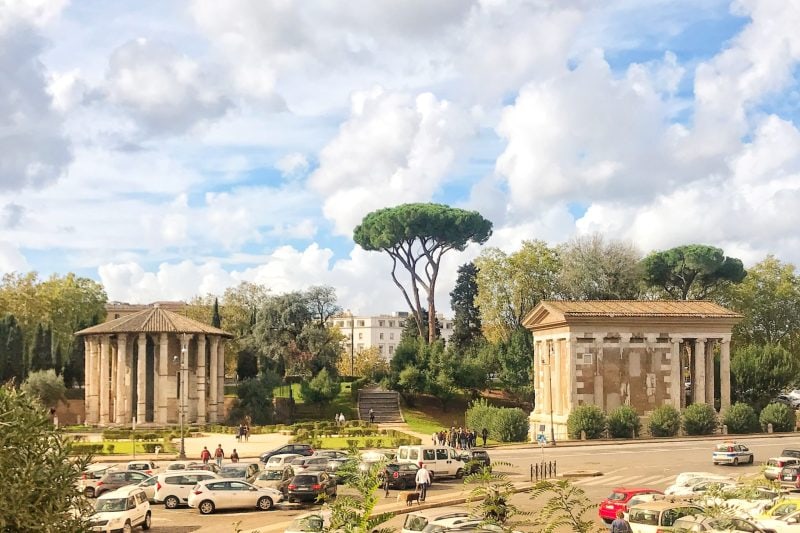Ancient stone temples in Rome near a road with parked cars and trees.