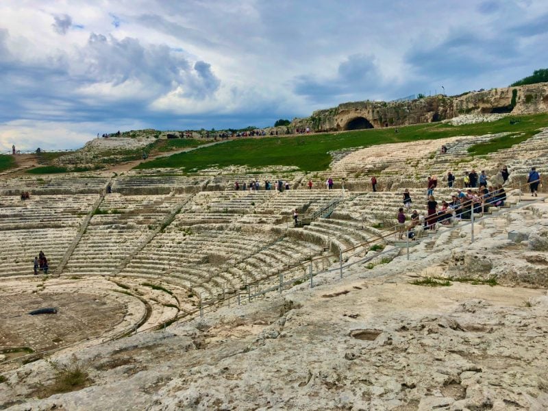Tourists walking through the ancient stone seating tiers of the Greek Theatre of Syracuse.