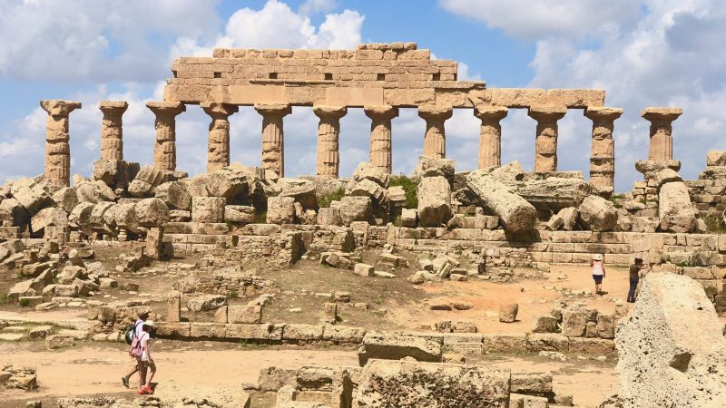 The ruins of an ancient Greek temple with a row of standing columns, with tourists walking through the site.
