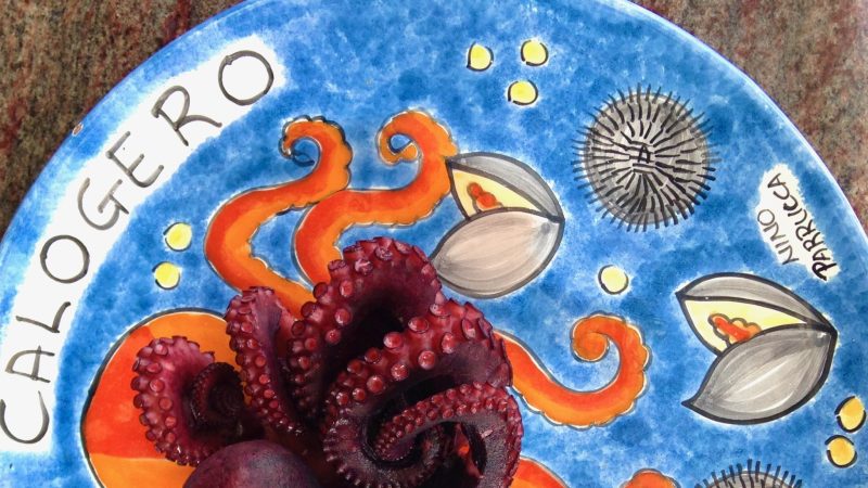 A cooked octopus tentacle served on a blue ceramic plate decorated with painted sea creatures and the name “Calogero.”