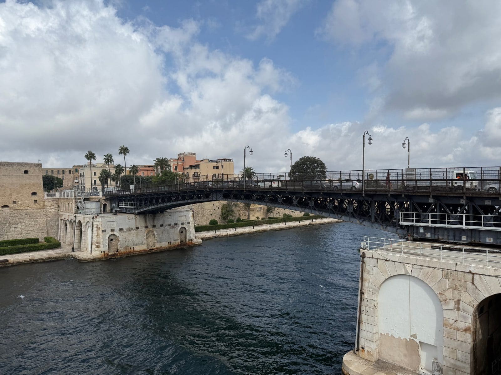 A metal swing bridge spanning a narrow waterway with buildings and stone fortifications on the shore.