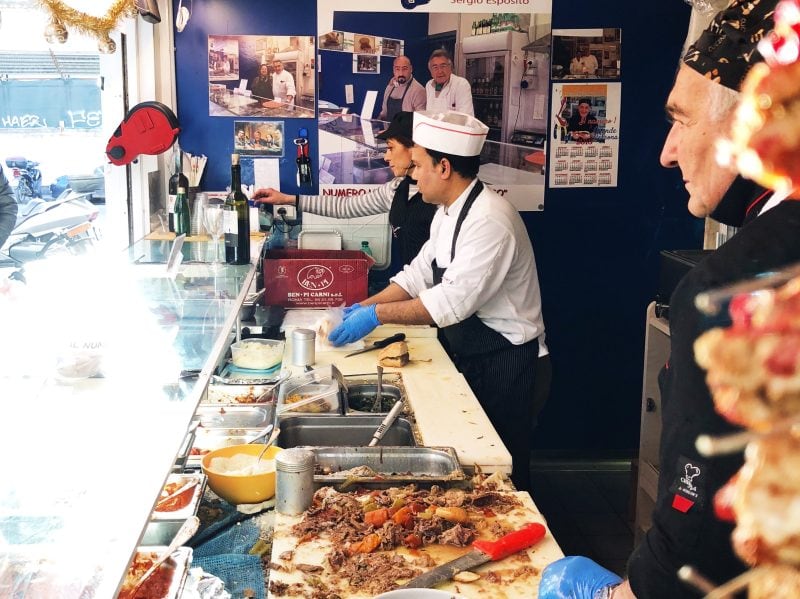 Three workers prepare food behind a deli counter with meat, condiments, and kitchen equipment visible; customers can be seen through the window. Posters hang on the wall behind the workers.