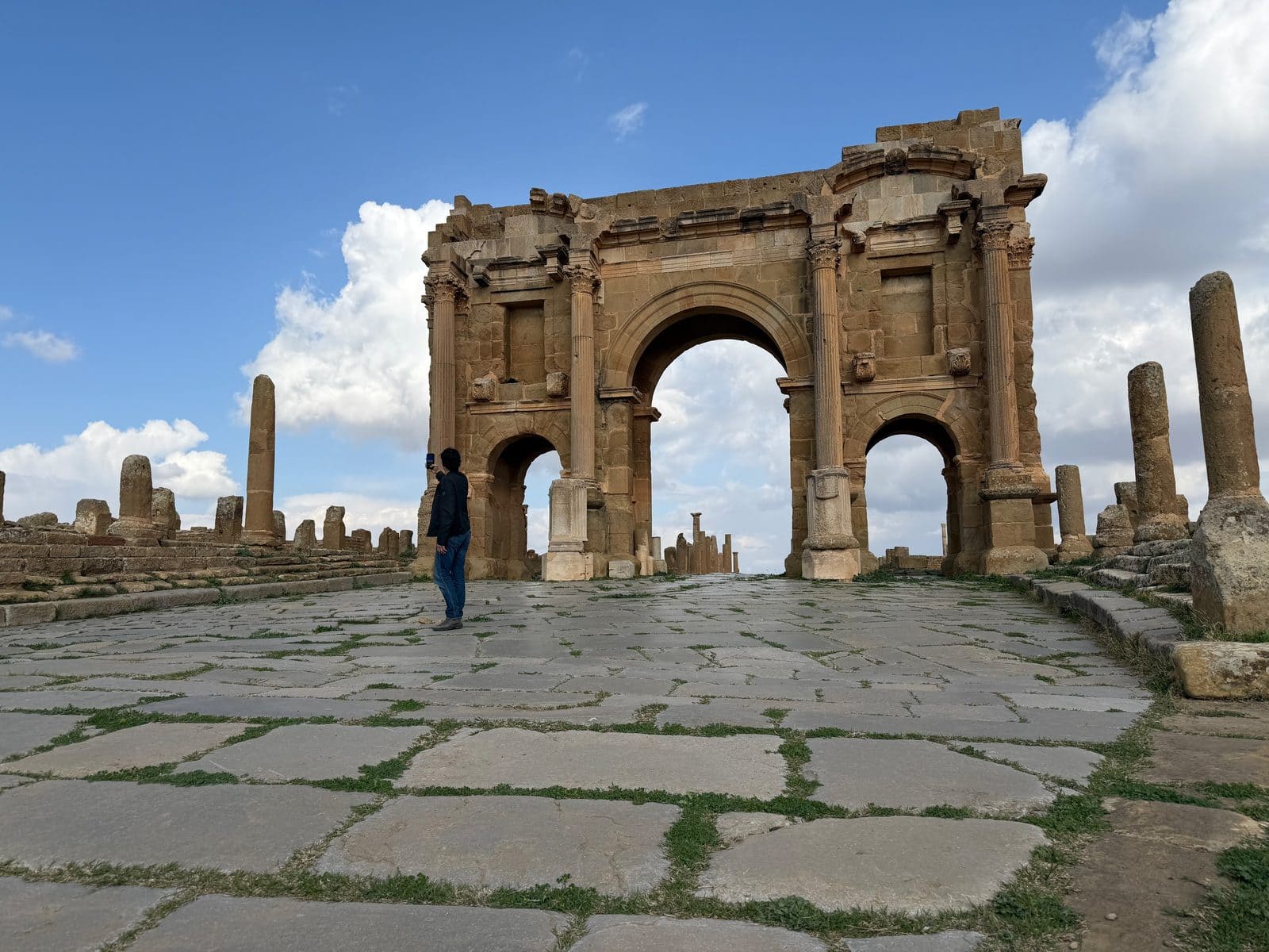 A person stands on a stone path taking a photo of a large ancient Roman triumphal arch.