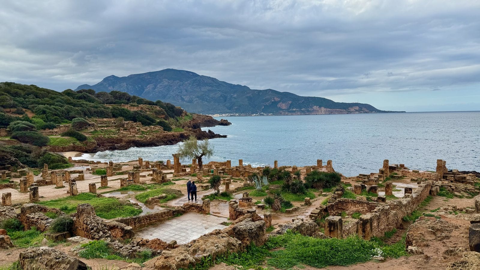 Ruins of the Roman city of Tipasa on the coast of Algeria, with two people standing near the columns overlooking the sea and mountains.