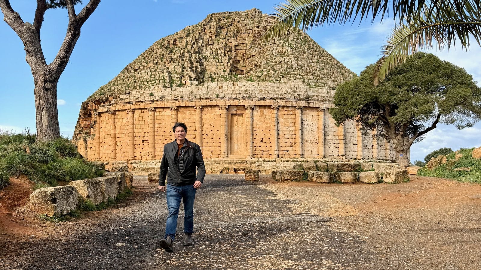 A man walks on a dirt path in front of the Royal Mausoleum of Mauretania, an ancient stone monument with a conical roof and colonnaded base.
