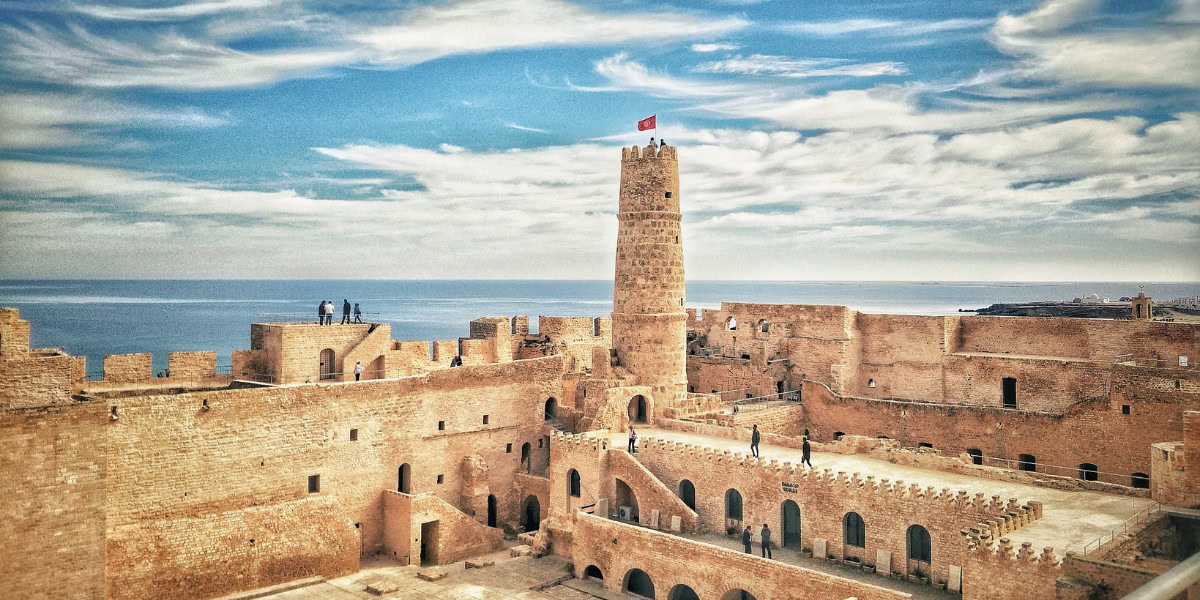 Ancient stone fortress by the sea with a tall tower flying a red flag, under a partly cloudy sky. A few people are visible on the walls and tower.