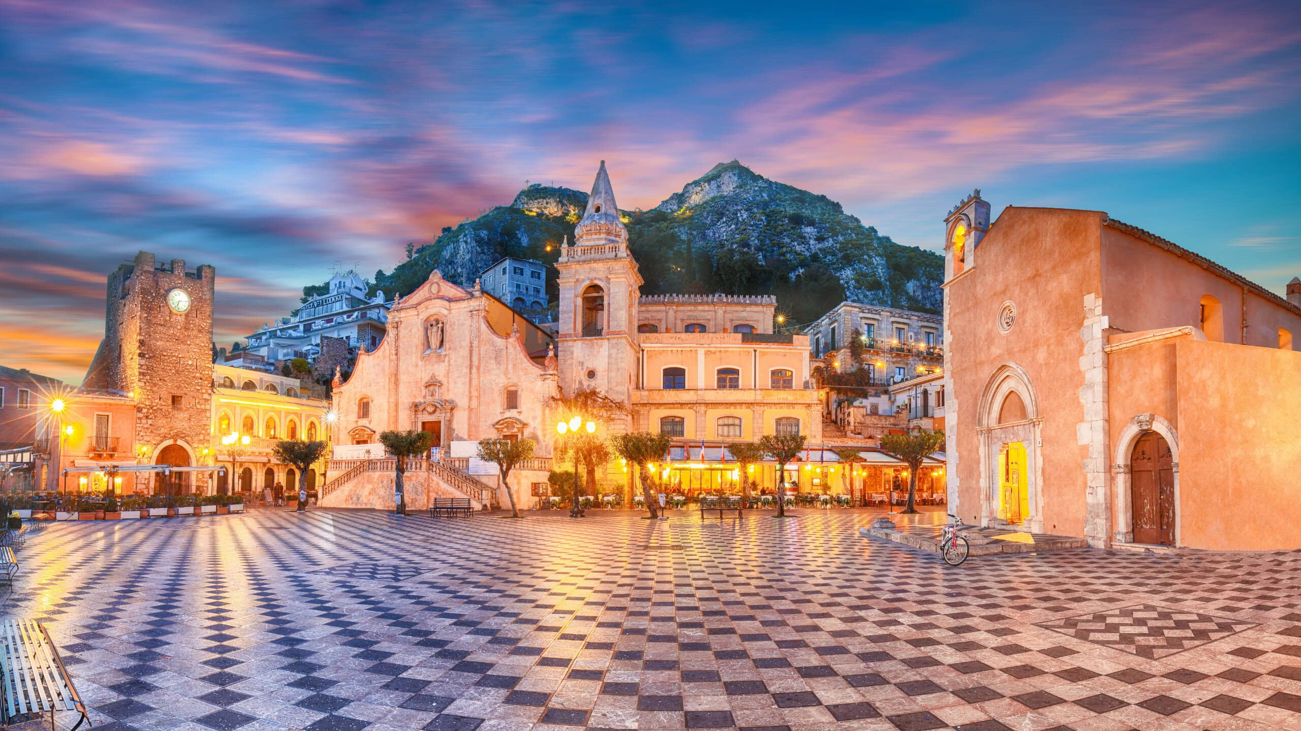 A historic town square with patterned stone tiles, illuminated buildings, and a mountain in the background at sunset.