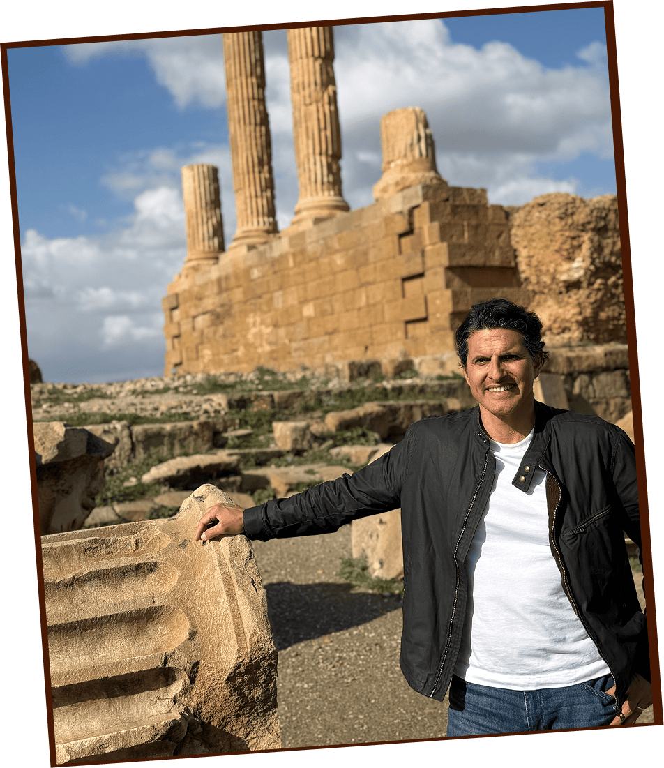 A man in a black jacket stands in front of ancient stone ruins with tall columns under a partly cloudy sky.