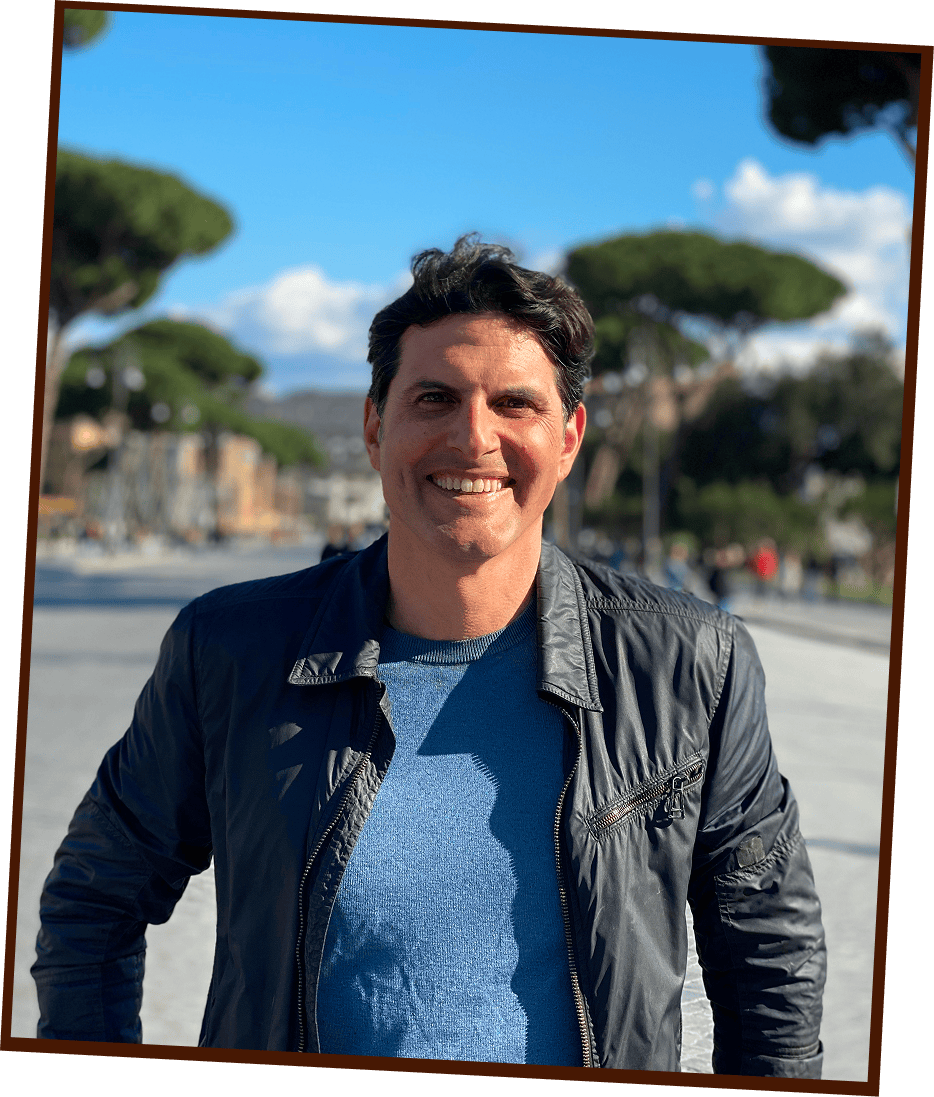 Man in a black jacket stands outdoors on a sunny day, smiling at the camera with trees and a blue sky in the background.