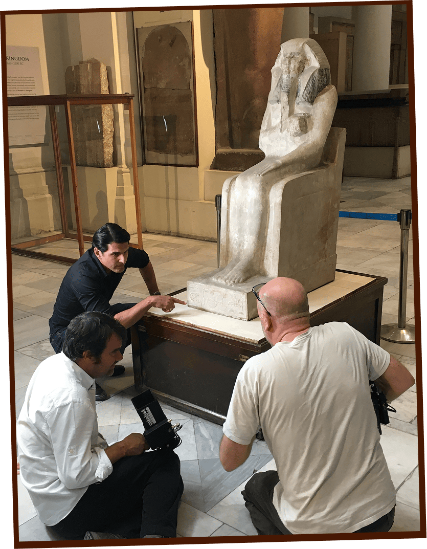 Three men examine and photograph a large seated stone statue inside a museum, focusing on details at the statue’s base.