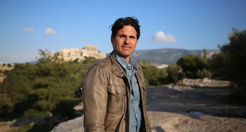 A man in a brown jacket stands outdoors with ancient ruins and a mountain in the background under a clear blue sky.