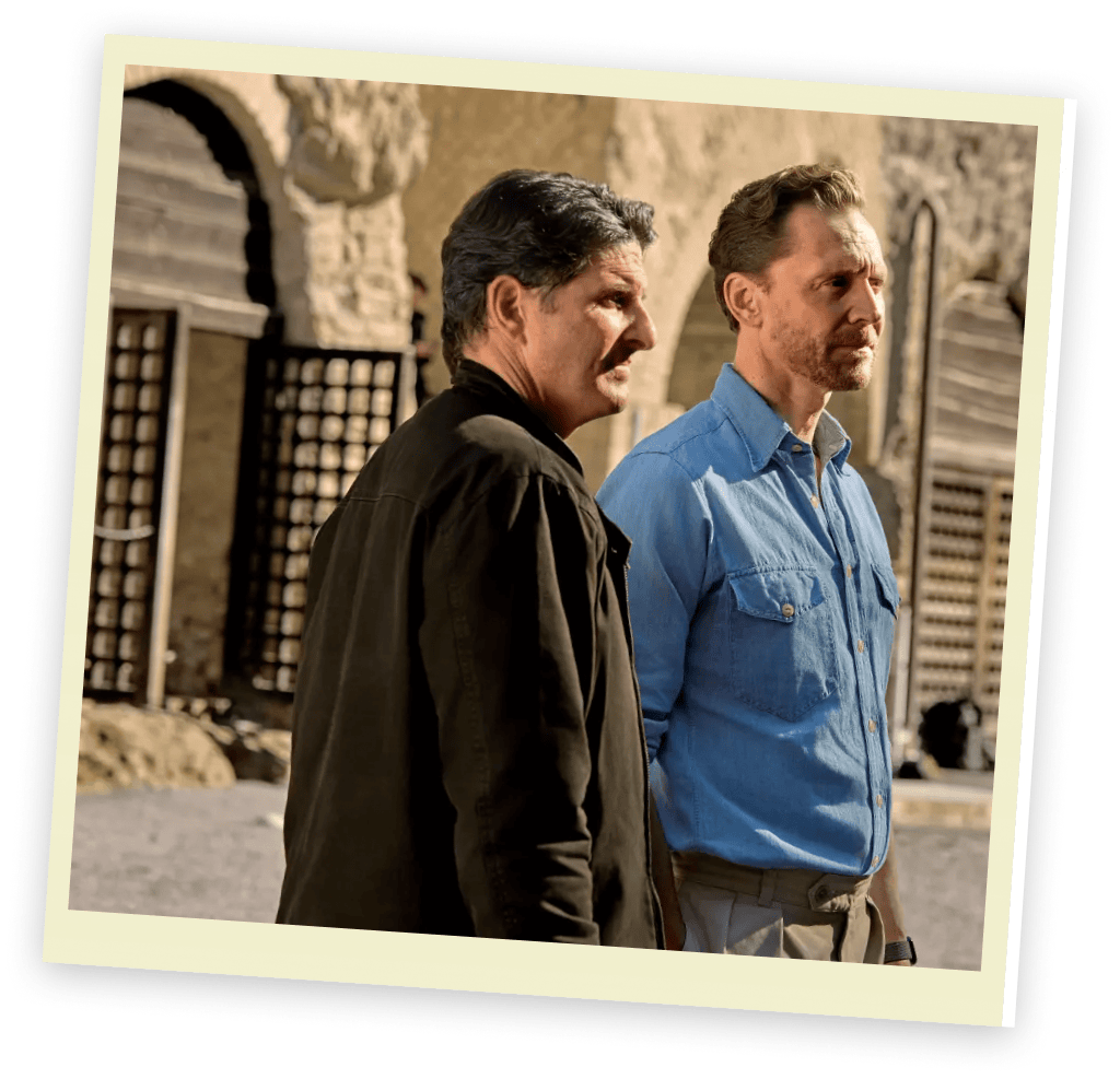 Two men stand outdoors in front of a stone building with wooden doors. One wears a dark jacket, the other a light blue shirt.