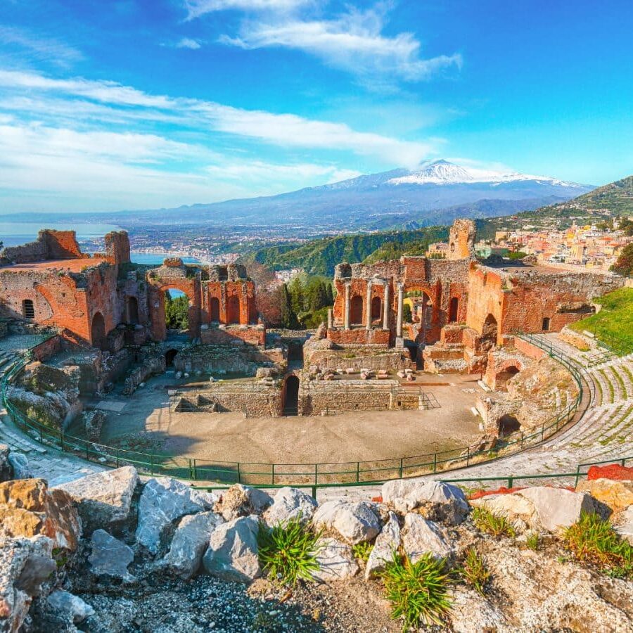 Ancient stone amphitheater ruins with tiered seating, surrounded by hills; a distant snow-capped mountain under a blue sky.