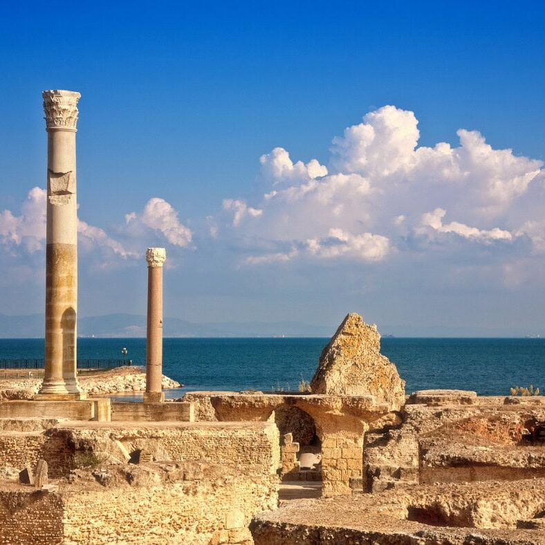 Ancient stone ruins with two standing columns overlook the sea under a blue sky with clouds.