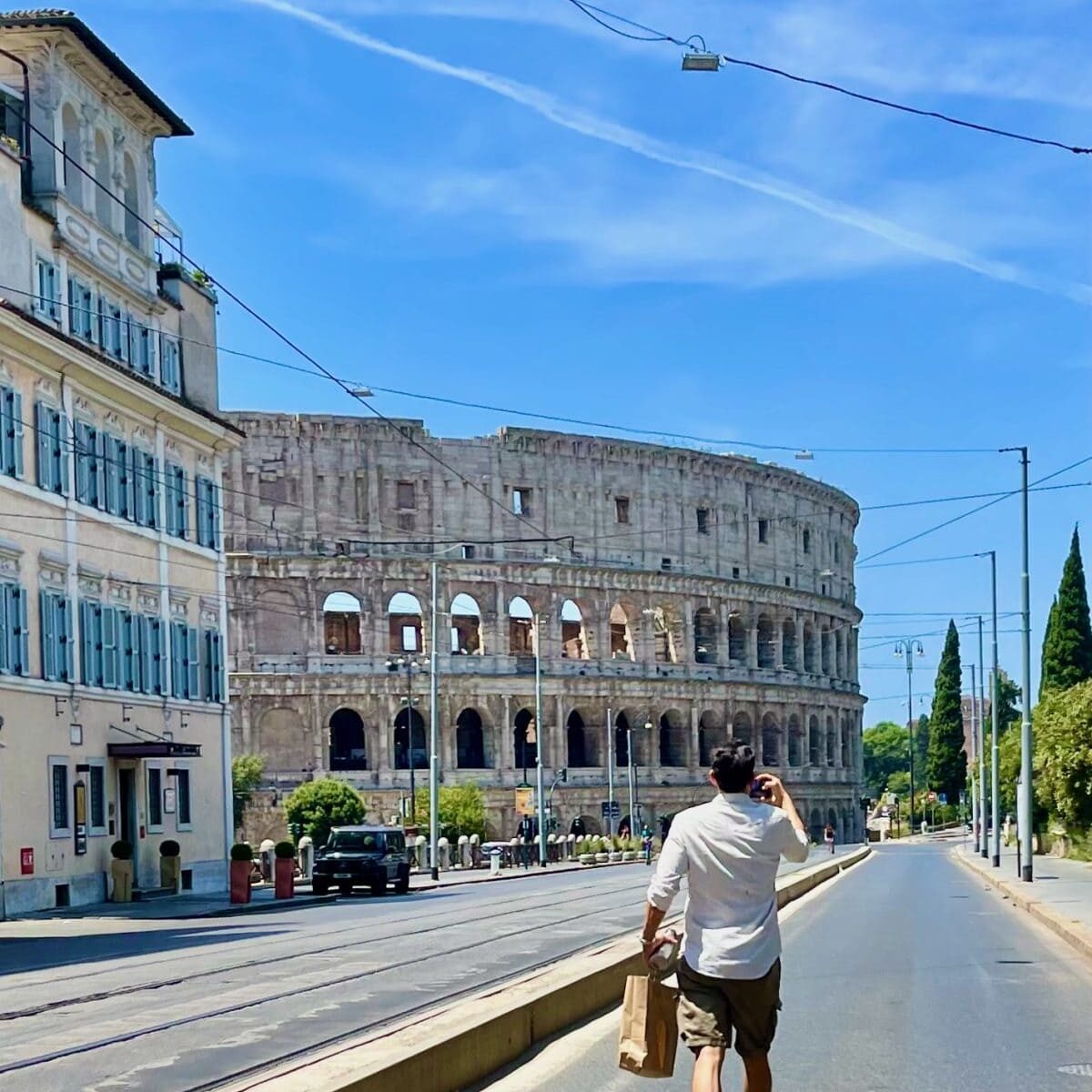 A man walks along a sunny street near the ancient Roman amphitheater in Nîmes, France, holding a paper bag and touching his head.
