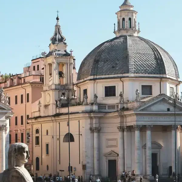 A large domed church with classical architecture stands in a busy city square with people walking and a stone statue in the foreground.