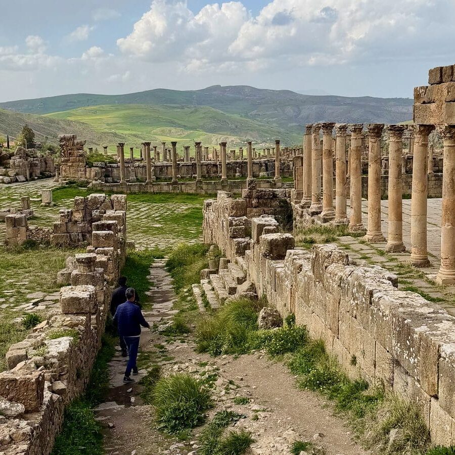 Two people walk through the ruins of the Roman city of Cuicul, featuring rows of standing stone columns and grassy courtyards under a cloudy sky.
