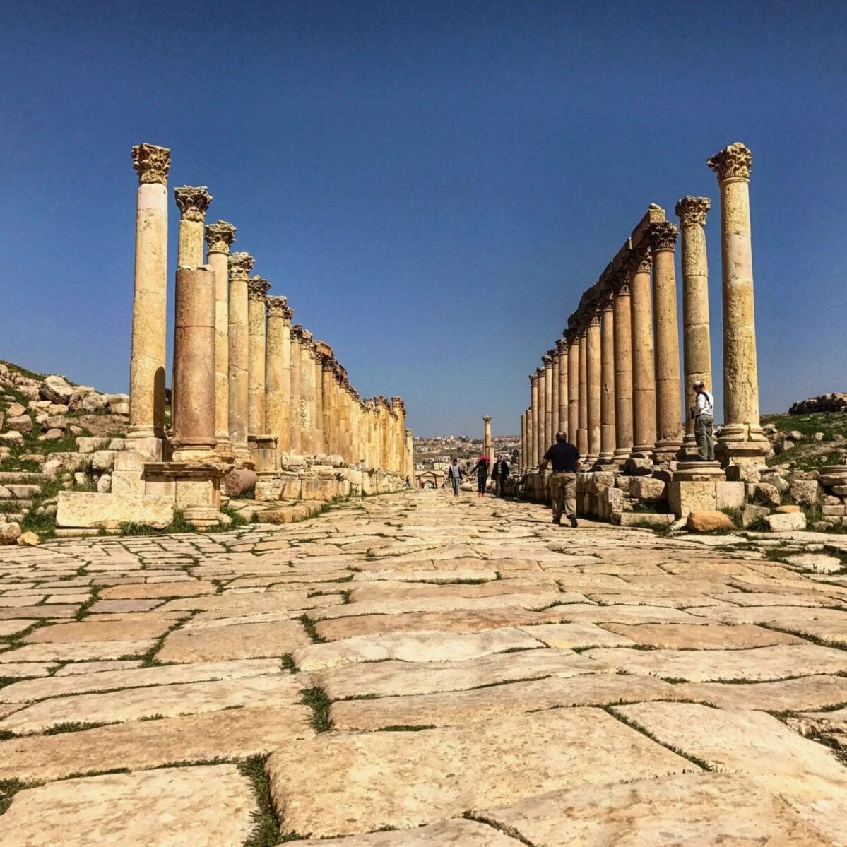 A wide, stone-paved street lined with rows of ancient Roman columns under a clear blue sky, with several people walking between them.