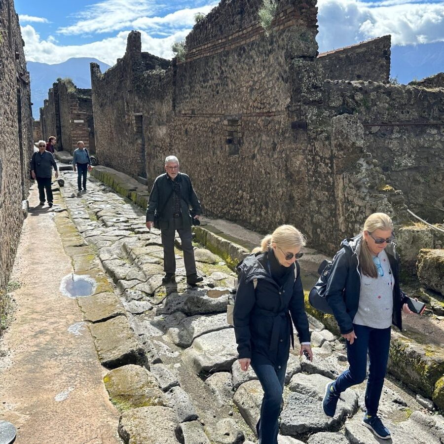 Tourists walk along a narrow, uneven stone street between ancient stone ruins in Pompeii.