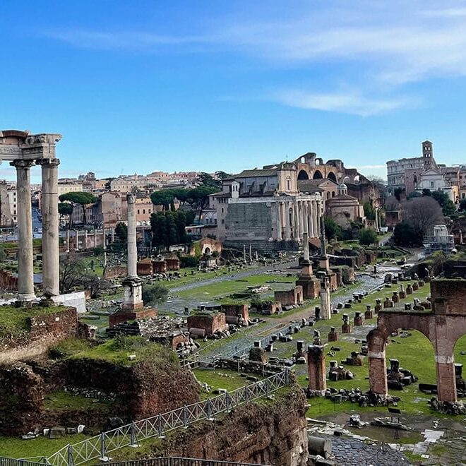 The Roman Forum, featuring ancient stone columns, ruins, and grassy pathways under a clear blue sky.