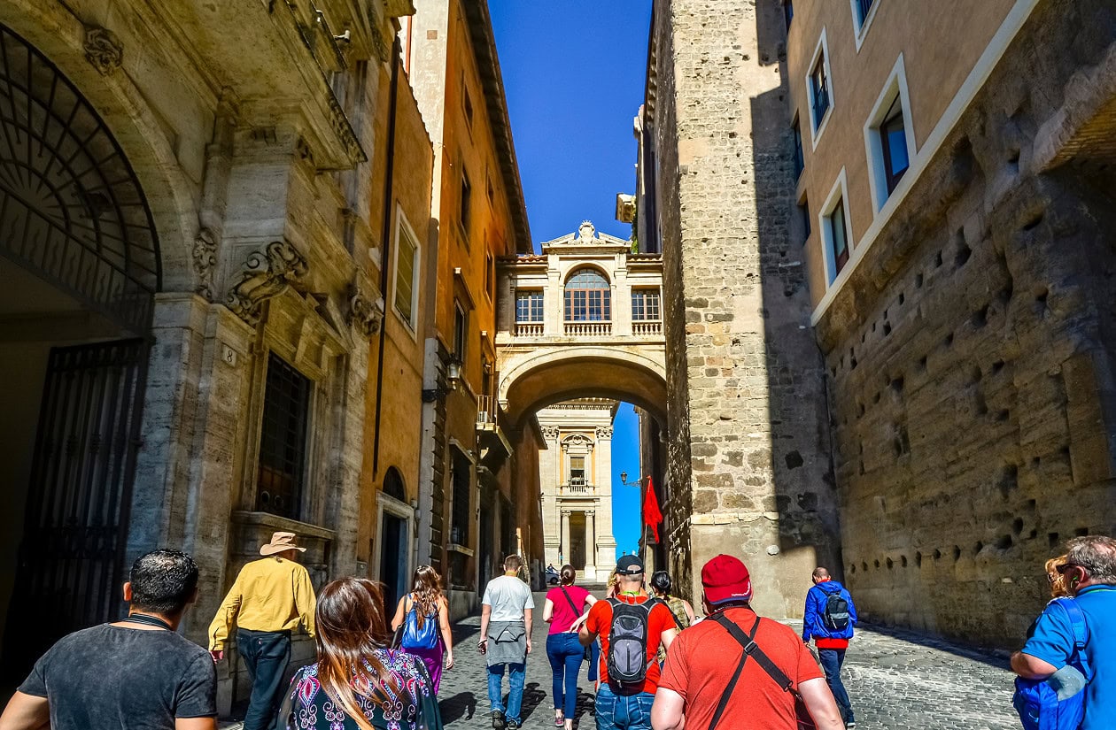 A group of tourists walks down a narrow cobblestone street in a historic European city, passing below an arched stone bridge connecting two buildings.