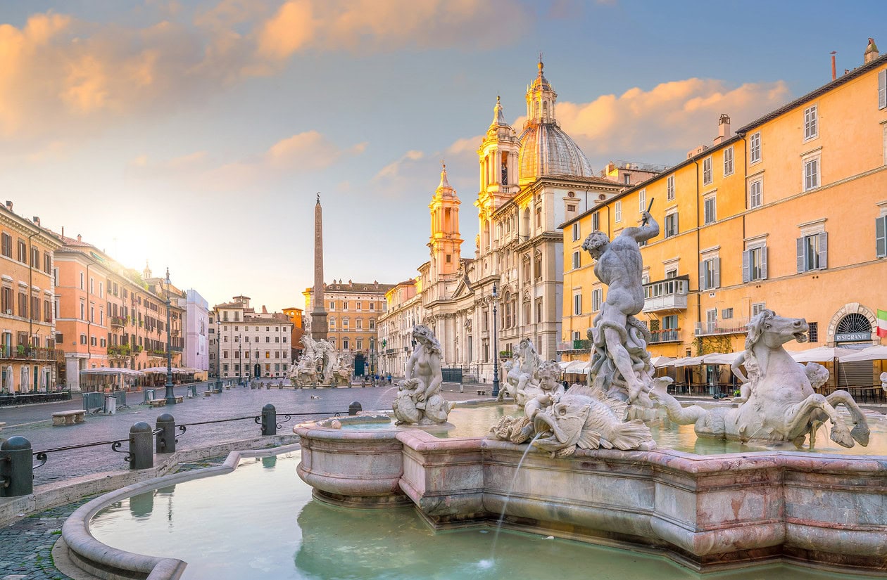 Piazza Navona in Rome at sunrise, featuring the Fountain of the Four Rivers, baroque buildings, and a clear sky with scattered clouds.