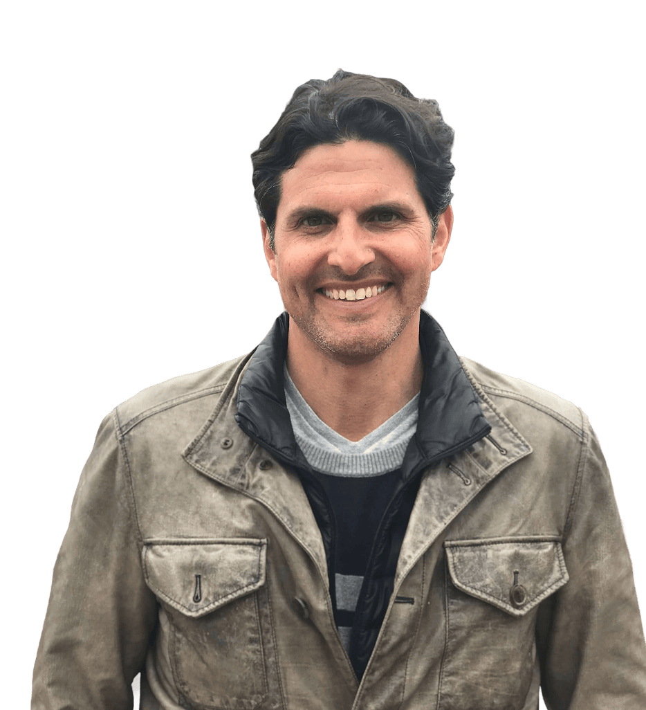 A man with dark hair is smiling at the camera, wearing a brown leather jacket over a black and gray shirt, with a plain white background.