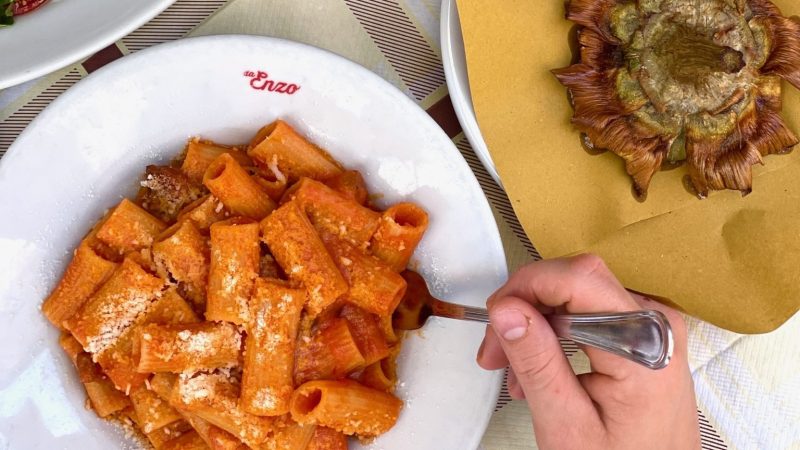 Rigatoni pasta with red sauce and grated cheese in a bowl labeled “da Enzo,” served with a fried artichoke.