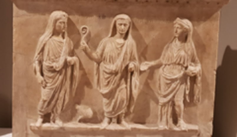 Stone relief showing three robed figures standing side by side, each facing forward, with draped clothing and distinct hand gestures.