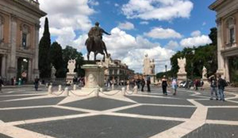 People walk around a large plaza with a central equestrian statue, surrounded by historic buildings and sculptures under a partly cloudy sky.