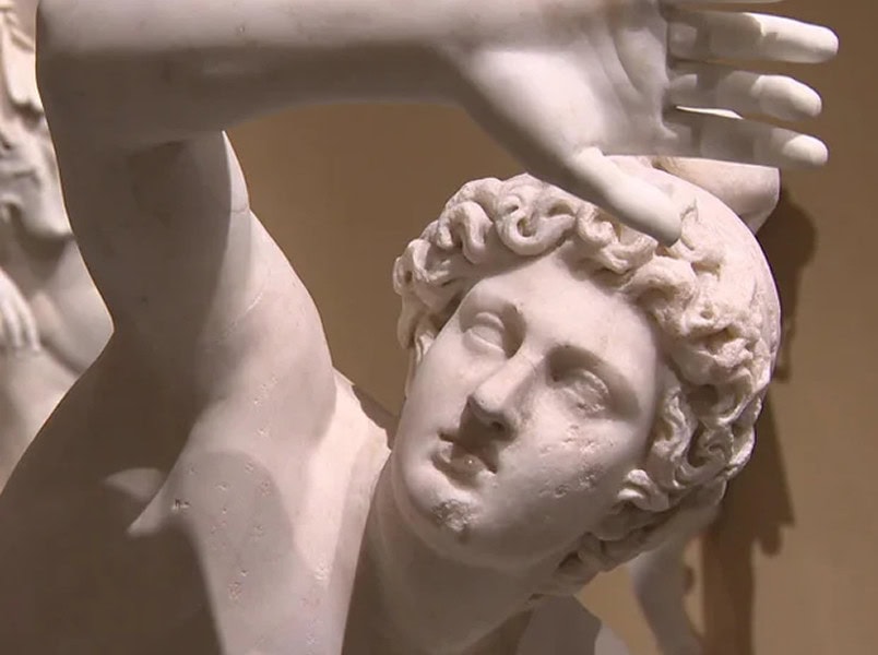 Marble statue of a figure with curly hair raising one arm and looking upward, showing detailed facial features and drapery.