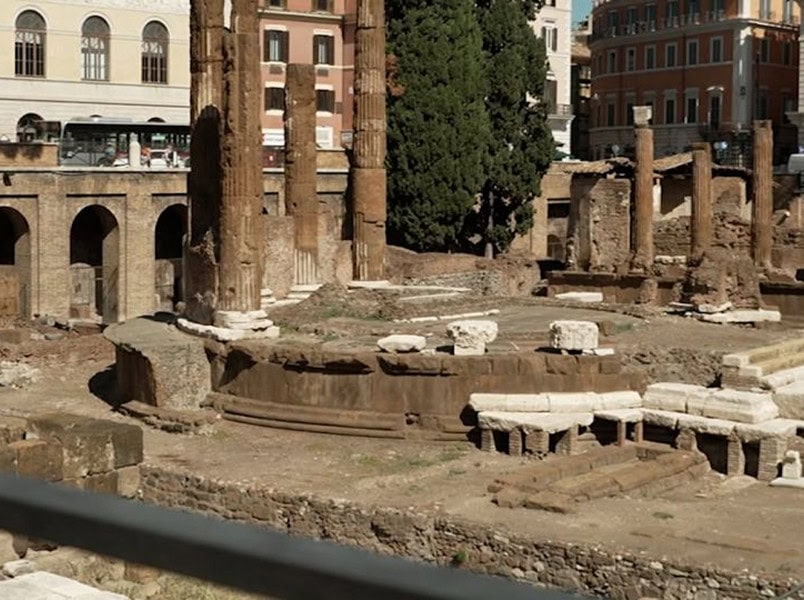 Ancient Roman ruins with stone columns and remnants of walls, surrounded by modern buildings and trees in the background.