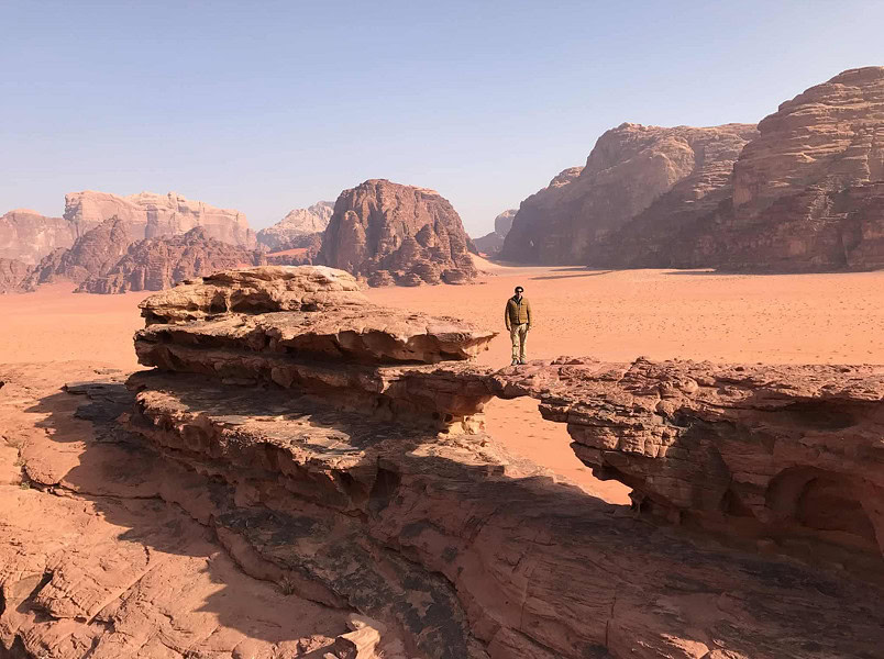 A person stands on a large rock formation in a vast, sandy desert surrounded by rugged, rocky mountains under a clear sky.
