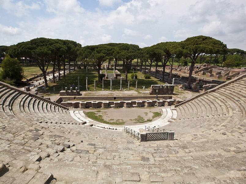 Ancient stone amphitheater with semi-circular seating and a central stage, surrounded by trees and ruins under a partly cloudy sky.