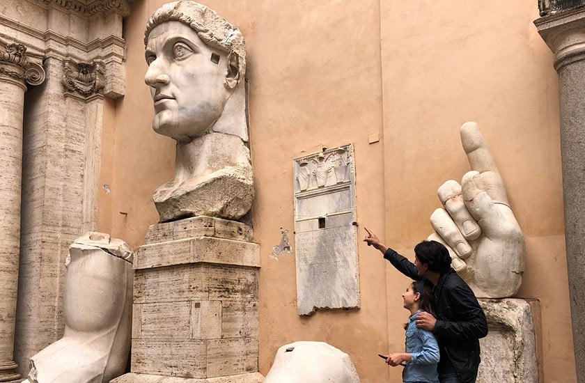 Two people look up and point at a large ancient marble head and hand sculpture displayed against a tan stone wall, with other statue fragments nearby.