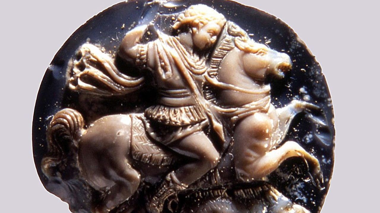 Ancient carved cameo showing a figure on horseback, wearing a tunic and raising an arm, with detailed horse and rider features against a dark background.