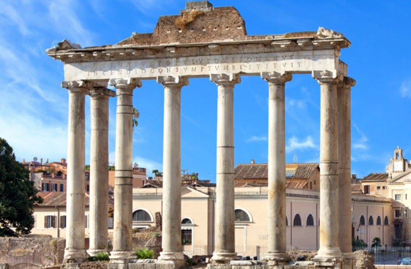Ruins of ancient Roman columns and a partially preserved building, set against a clear blue sky with some city structures in the background.