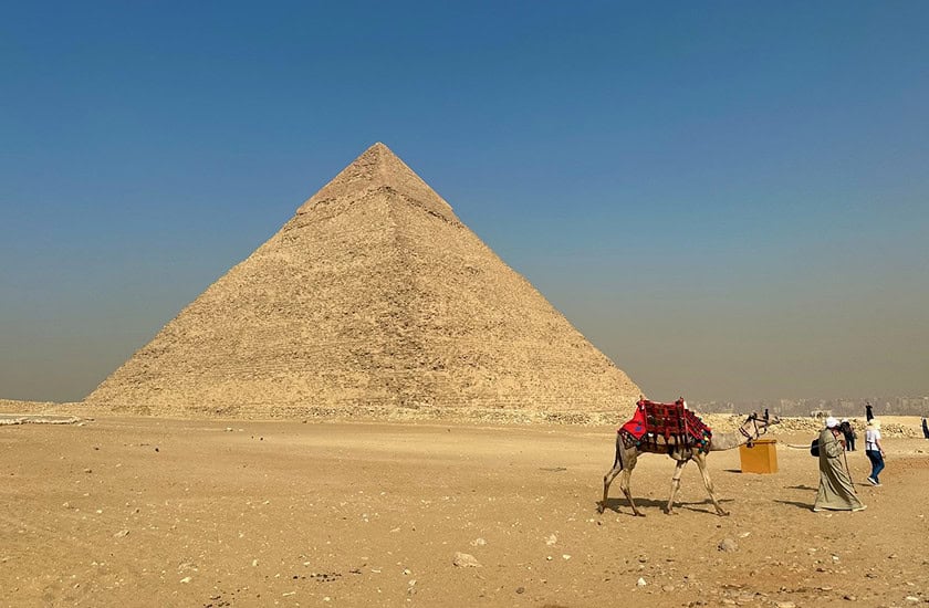 A large pyramid stands in a desert under a clear sky. In the foreground, two people lead two camels covered with red blankets.
